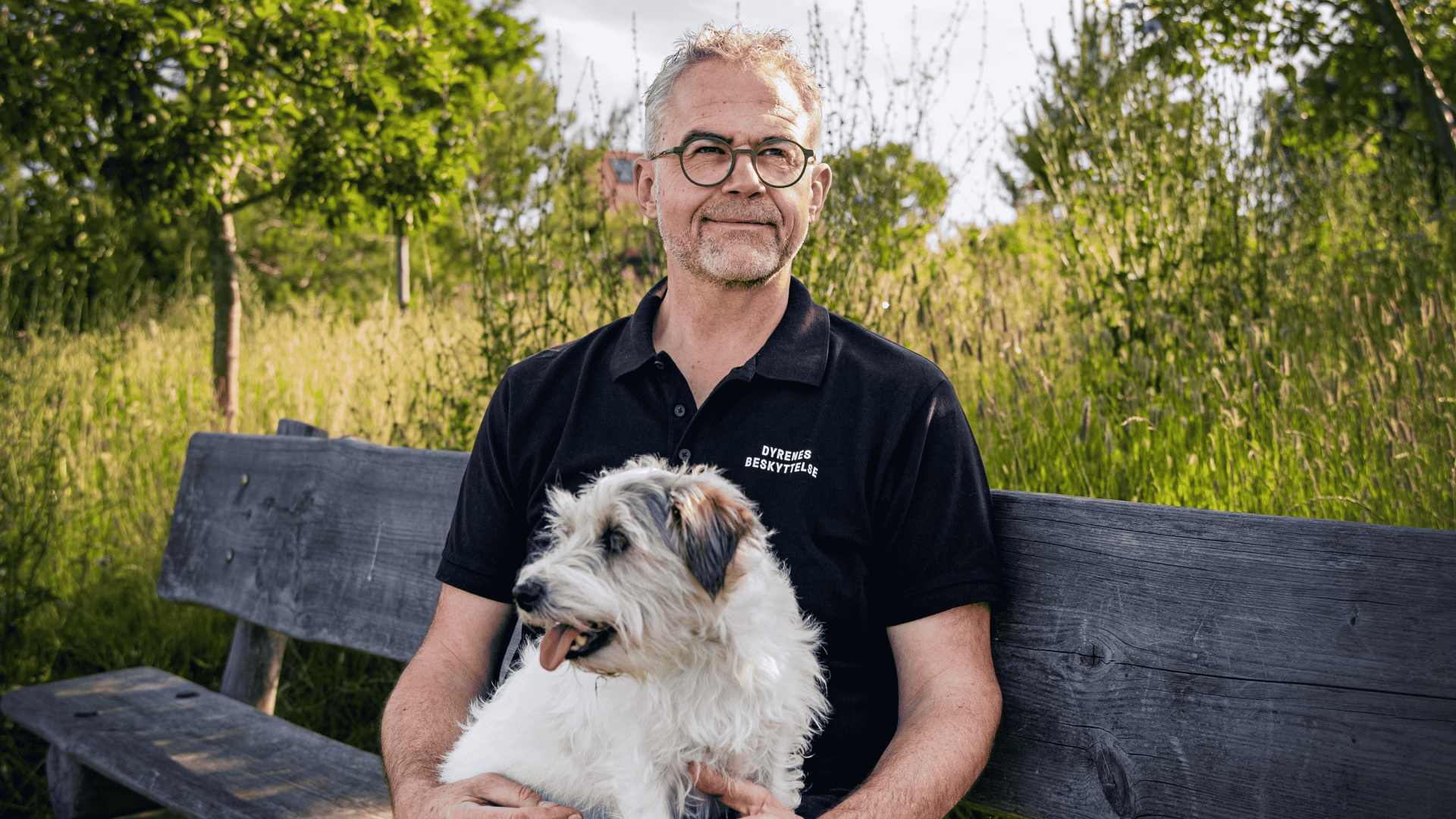 Jens Jokumsen sidder på en bænk i græsset med sin hund i skødet