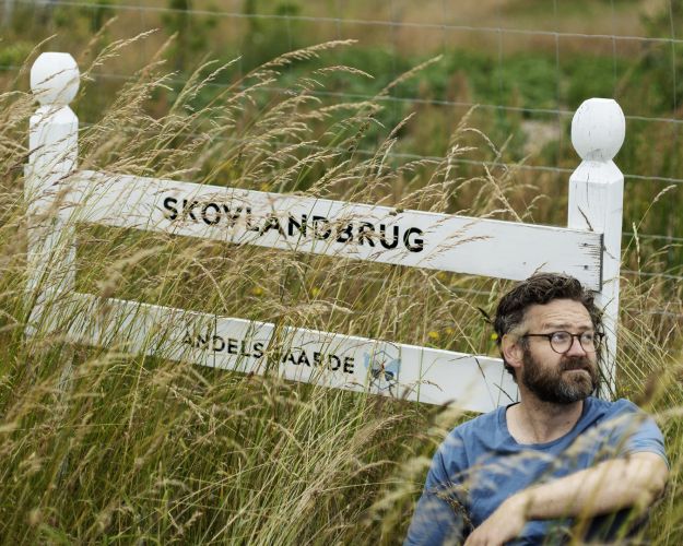 Rasmus Willig sidder i højt græs ved et skilt, hvor der står 'Skovlandbrug'